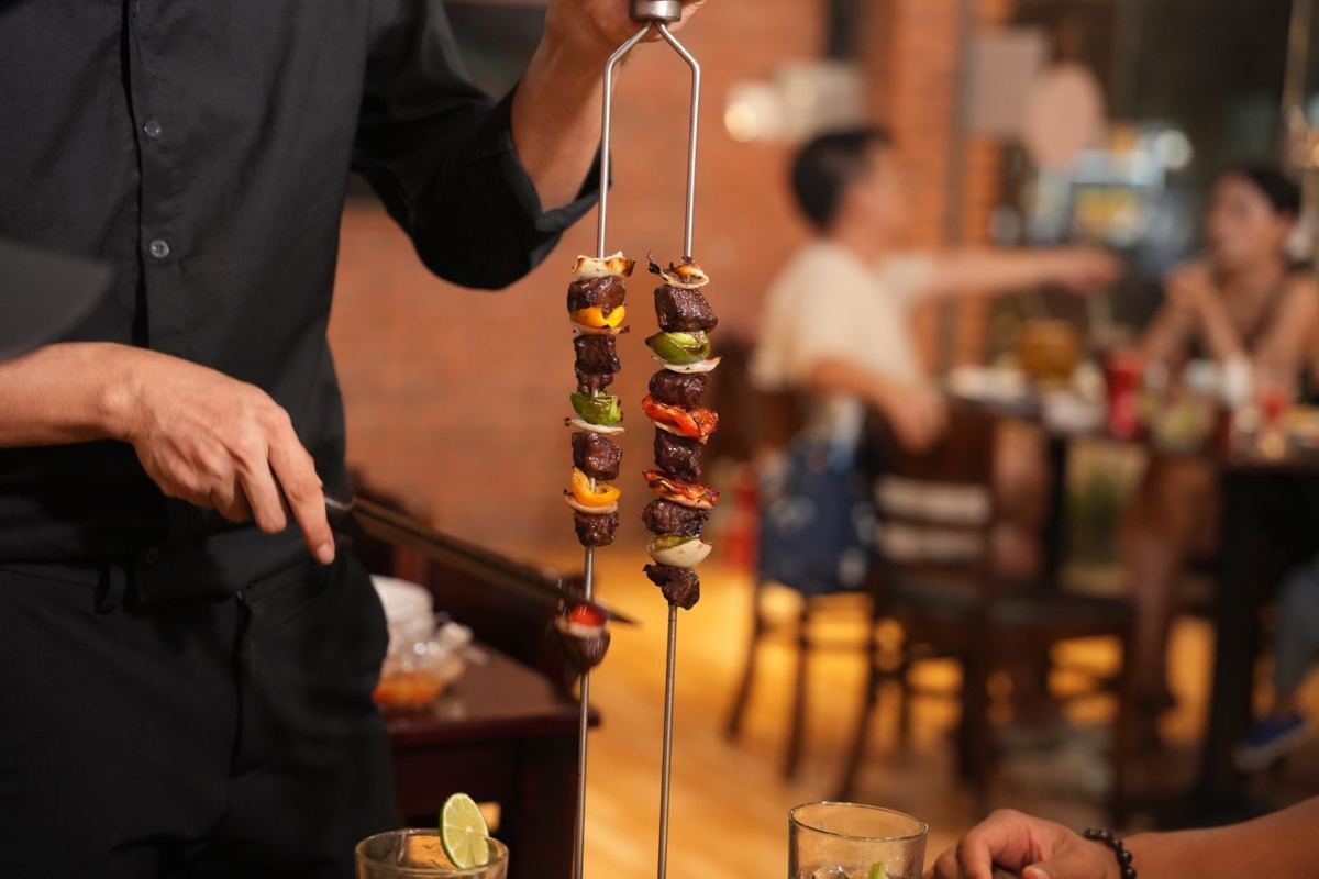 A passador holding a skewer of grilled meat and vegetables at Brasa Brazilian BBQ